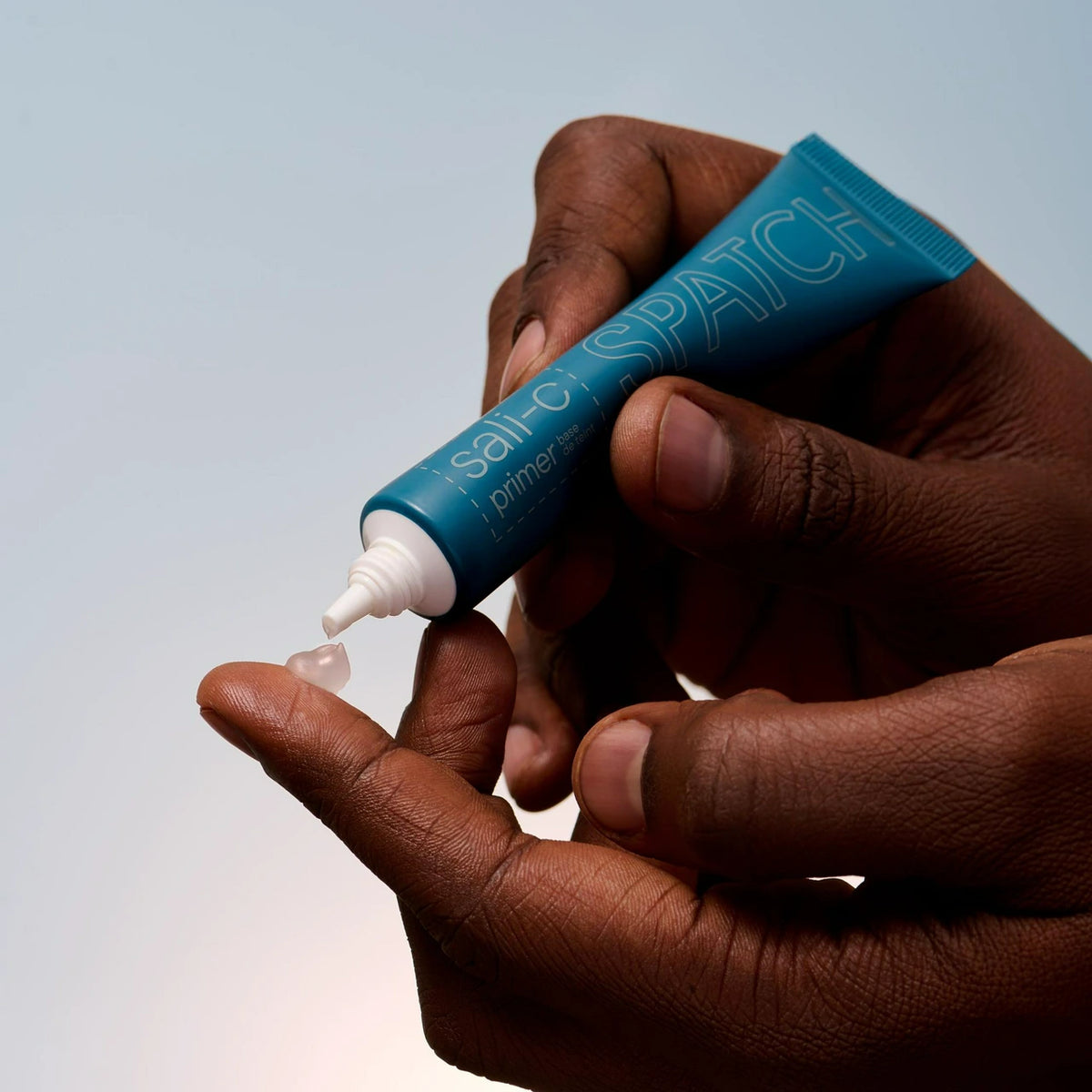 A close-up of a person's hands squeezing a teal tube of Spatch primer onto their finger, with a small clear gel-like droplet falling, against a soft light gray background.
