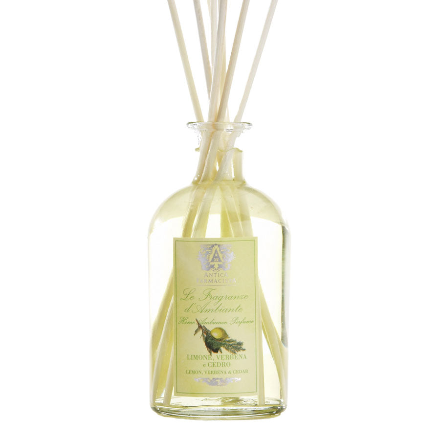 A clear glass diffuser bottle with several white reeds inserted, and a label that reads 'L'Essenza del Mediterraneo' with an image of a green leaf.