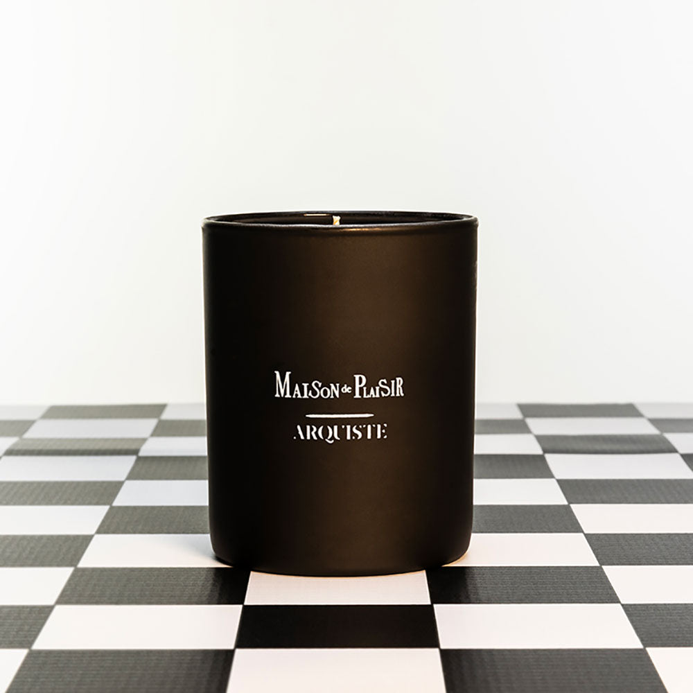 A black candle with a white label reading 'MAISON DE PLAISIR ARQUISTE' sitting on a checkered surface