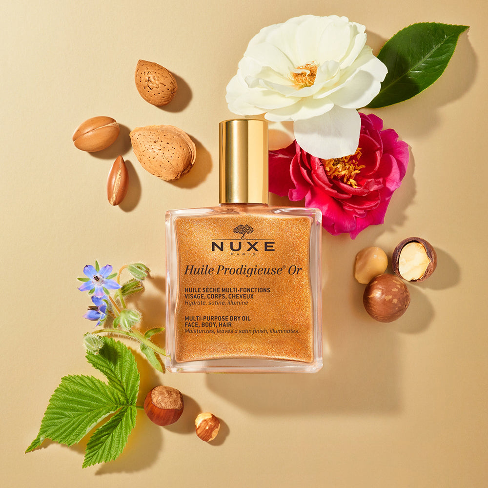 Nuxe Huile Prodigieuse Or on a tan background surrounded by botanicals. 