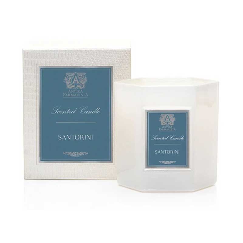 A white hexagonal candle jar with a blue label that reads 'SANTORINI' on the front, indicating the fragrance of the candle, next to its box.