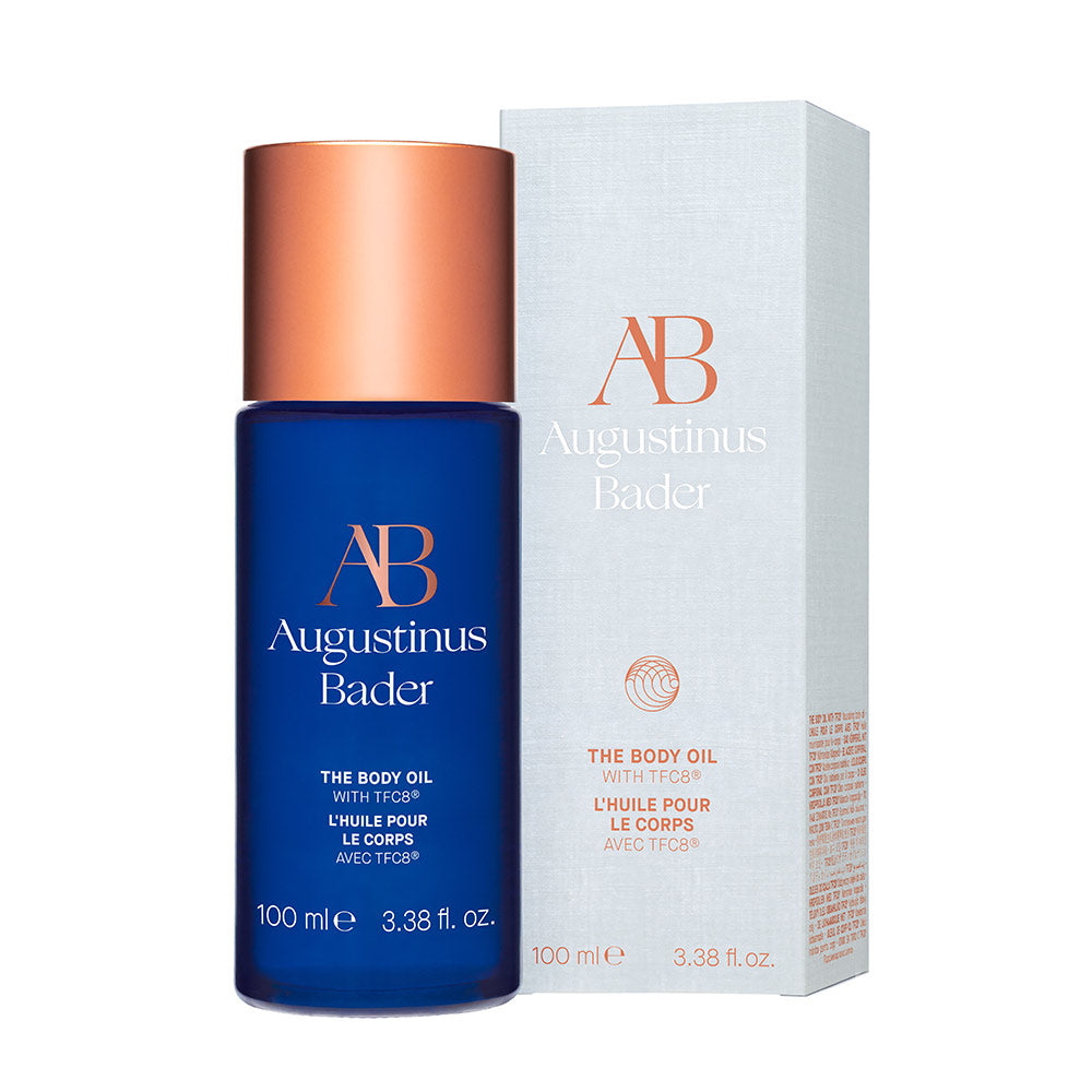 A bottle of Augustinus Bader The Body Oil with blue and copper packaging, labeled 'AB' for Augustinus Bader.