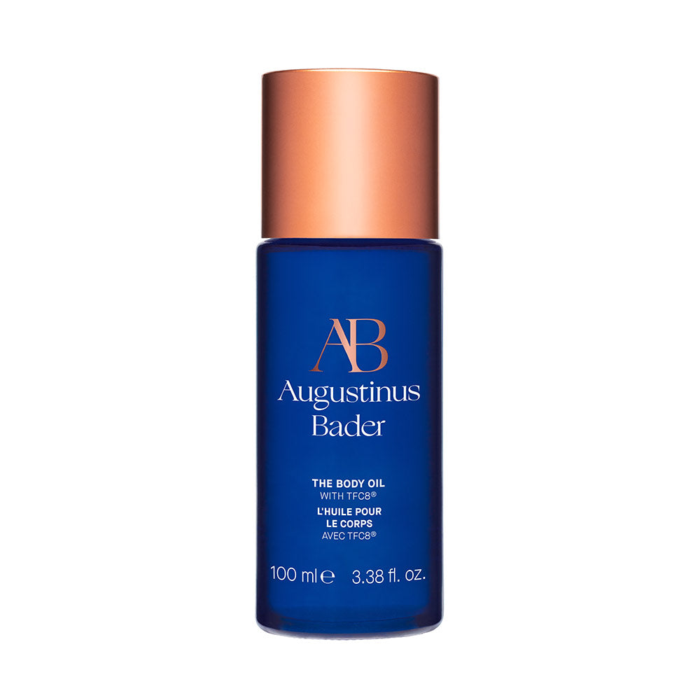 A bottle of Augustinus Bader The Body Oil with blue and copper packaging, labeled 'AB' for Augustinus Bader.