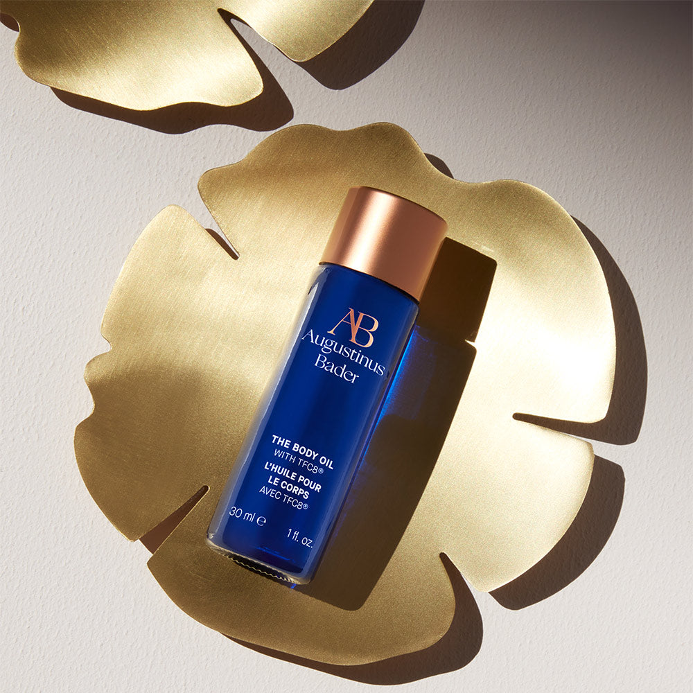 A bottle of Augustinus Bader The Body Oil with blue and copper packaging on a golden plate
