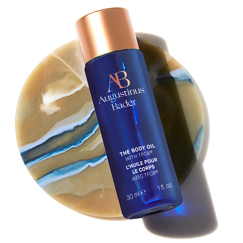 A bottle of Augustinus Bader The Body Oil with blue and copper packaging on a circular marble object