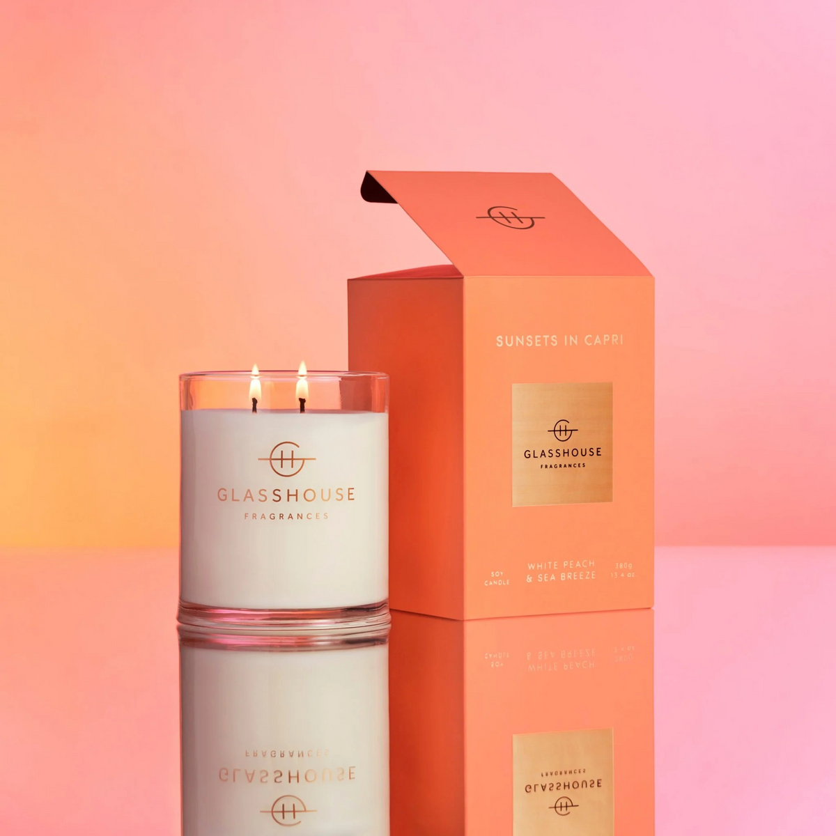 Glasshouse Sunsets in Capri Candle next to it's orange outer-packaging box on a reflective surface infront of pink and orange ombre background.